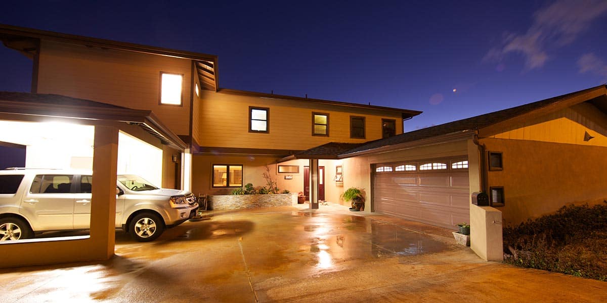Two-story Oahu house at dusk with lights on, a wet driveway, an SUV parked in an open garage, and a closed double garage door—perfect for future additions or expansion.