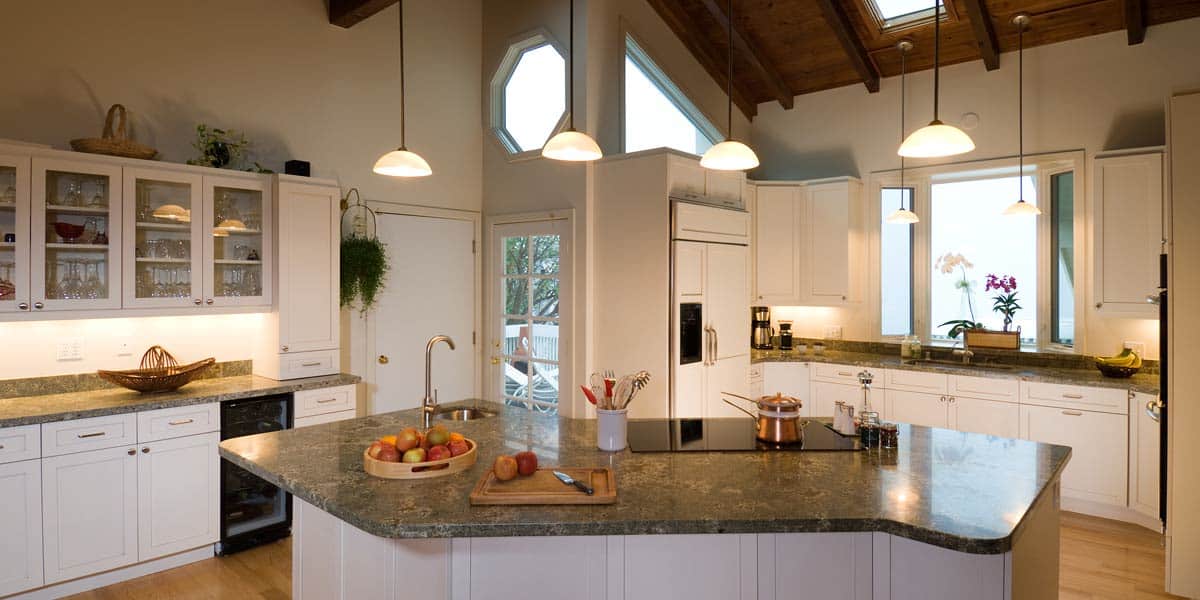 Modern kitchen by Oahu Contractor Services with white cabinets, green marble countertops, pendant lights, and a central island featuring fruit, cutting board, and cooking pots. Large windows and skylights flood the space with natural light.