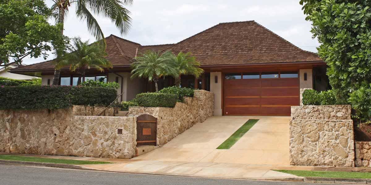 Single-story house with a dark wood shingle roof, stone walls, a wooden gate, and a two-car garage, surrounded by palm trees and tropical plants—crafted with quality by Oahu Contractor Services.